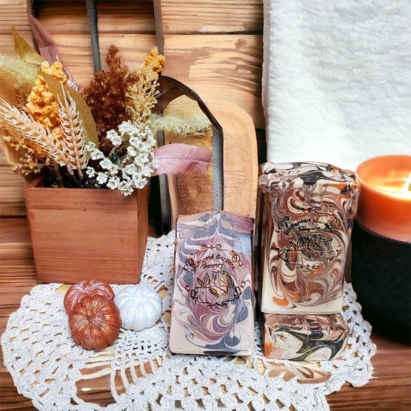 Two bars of handmade soap with floral and pumpkin patterns, placed on a doily beside a candle and a basket of decorative items.