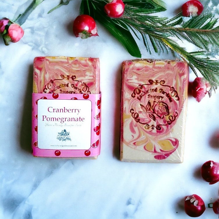 Two bars of handmade soap with 'Cranberry Pomegranate' label, surrounded by decorative elements like pink and red berries, green leaves, and a sprig of dried flowers.