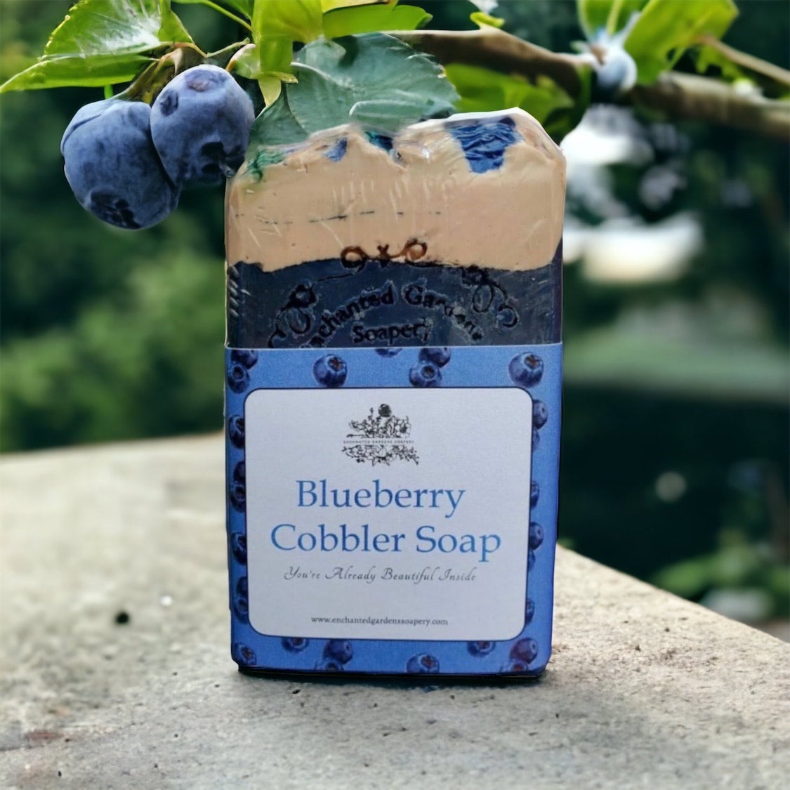 A bar of Blueberry Cobbler Soap with a label that includes the scent marked as blueberry, and a bundle of blueberries in the background, placed on a stone surface with greenery in the background.