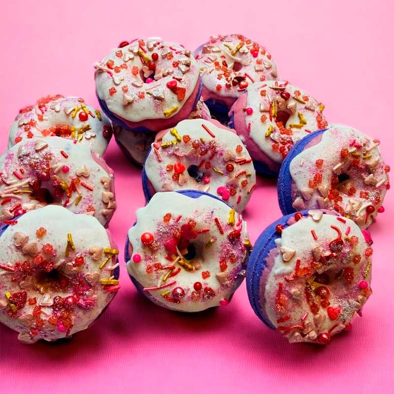 A collection of bath bombs arranged on a pink background, each shaped like a ring with a variety of colorful sprinkles on top.