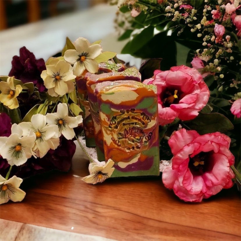 A colorful bar of Gardenia Luxury Soap with layers of green, pink, dark orange, and white, displayed among various flowers.