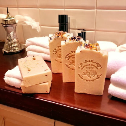 A selection of handmade bar soaps with a light color, patterned with floral and botanical designs, displayed on a counter with a candle and towels in the background.